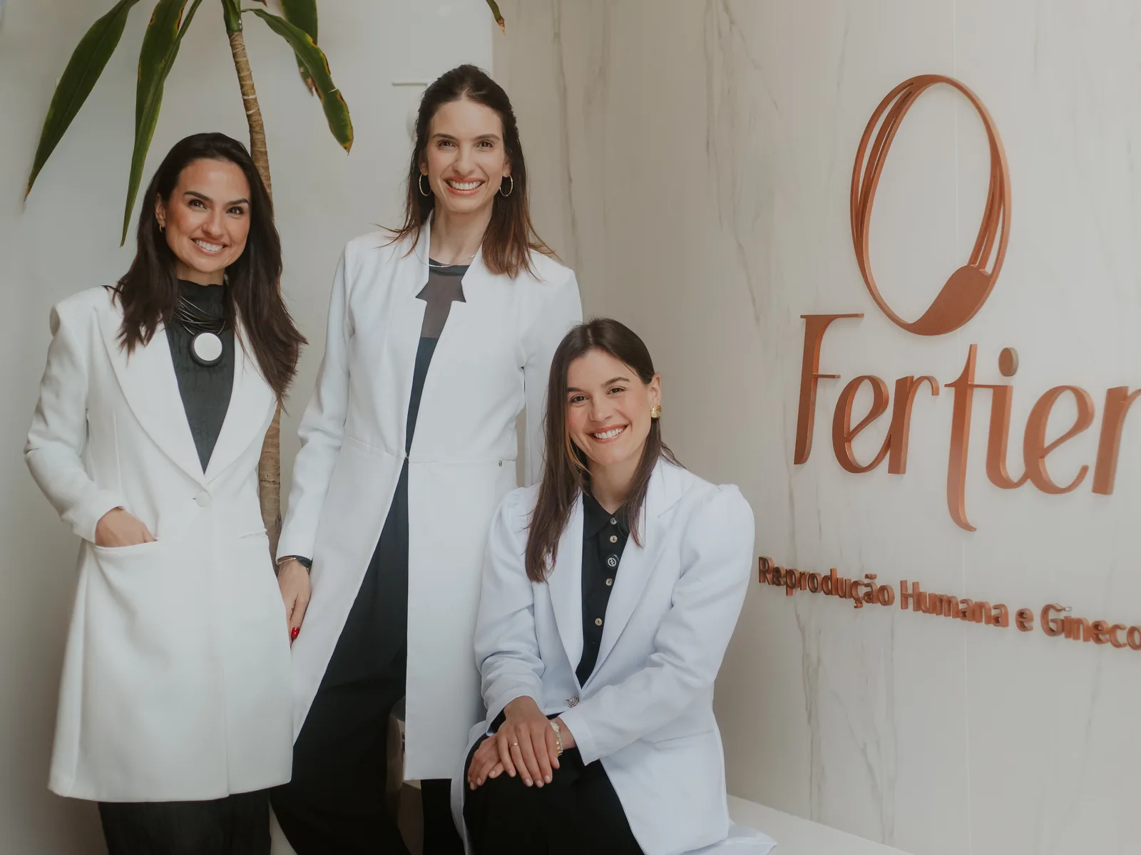 As três médicas da Clínica Fertier diante da parede com a marca da clínica
